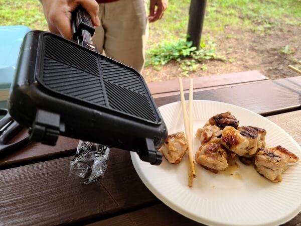 Someone holding a closed hot sandwich press, next to a plate of cooked chicken pieces set on a brown picnic table. There is a pair of chopsticks on the plate next to the chicken.