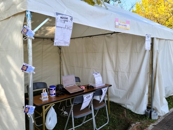 A white outdoor event tent booth with a laptop, speakers, and flyers on a table. In the foreground are Nice Gear Games business cards taped to a tentpole, and a Nice Gear Games sign over the tent. There are also flyers for 同ZINE hanging down from the tent and off the backs of the chairs.