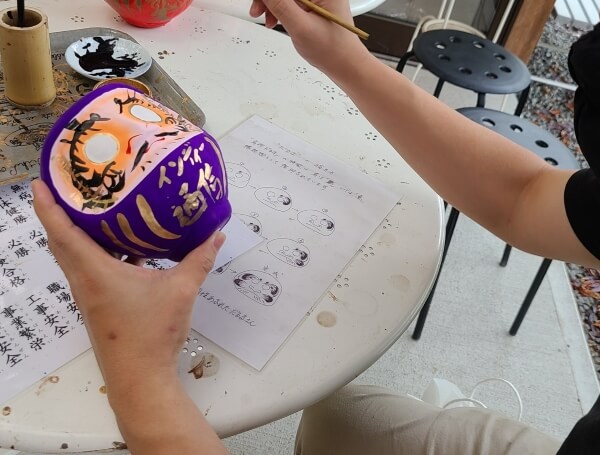 Person holding a purple daruma in one hand and paintbrush in the other. Daruma has gold letters down the front reading インディー通信 Indie Tsushin.