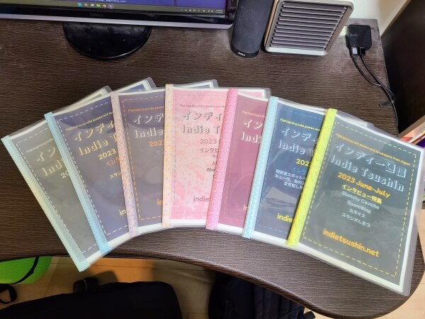 Seven colored print-out issues of Indie Tsushin fanned out on a desk.