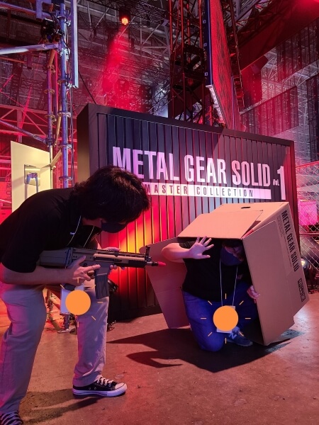 Person holding a toy gun and lifting up a cardboard box, underneath which is another person holding up their hands in surrender. In the background is a metal shipping container reading METAL GEAR SOLID Vol 1: Master Collection