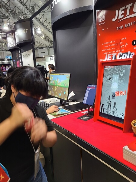 Person shaking a bottle of cola in front of the Jet Cola booth at TGS