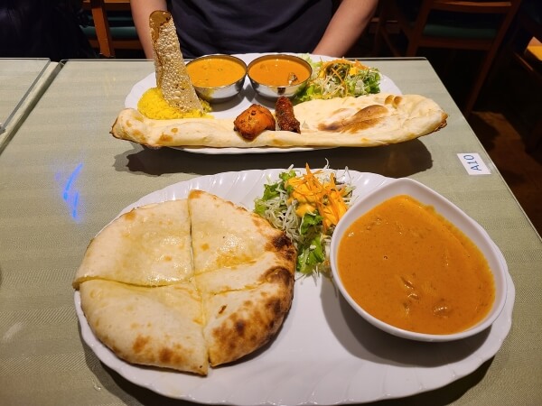 A plate of curry with a circular cheese naan cut into fourths and a bowl of butter curry next to it. Across the table is a long piece of naan, two pieces of chicken, two small bowls of curry, and a cylinder of fried naan.